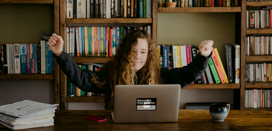 Child cheering behind a laptop sitting in front of a bookshelf
