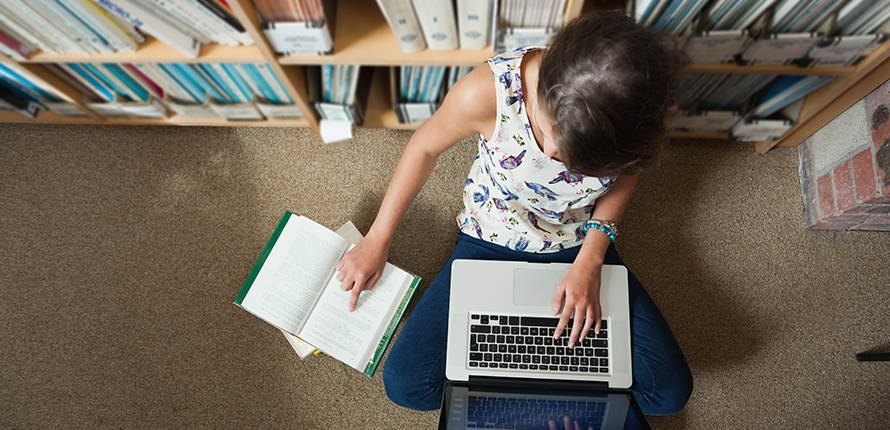 Person sitting on the floor in front of a bookshelf reading a book and typing on a laptop
